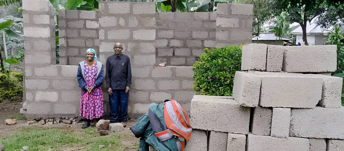 An elderly couple, with the woman wearing a vibrant patterned dress and the man in a dark shirt, stand in front of an unfinished brick structure surrounded by greenery. Nearby, there are stacks of concrete bricks.