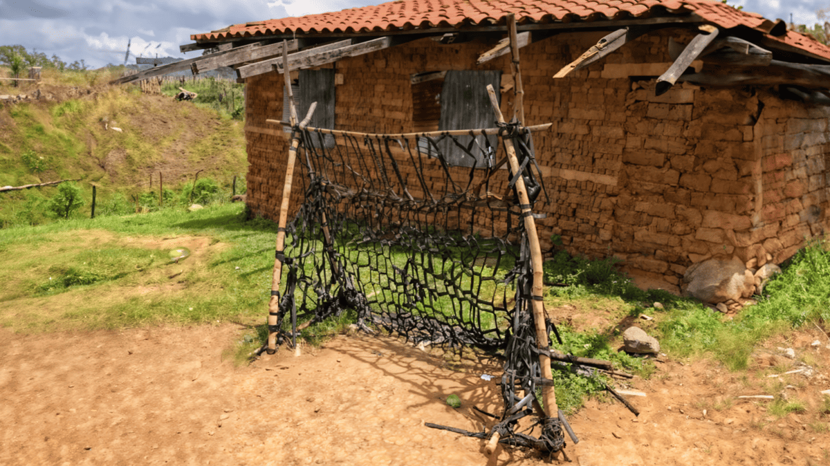 A makeshift soccer goal created ingeniously from discarded black irrigation tubes woven tightly across a wooden frame. Set against the backdrop of a brick house in a rural setting, the goal symbolizes the innovation and resourcefulness of local Honduran children, transforming agricultural waste into a playfield essential. The ground, compacted earth, bears evidence of frequent use, showcasing the passion and dreams of young football enthusiasts.