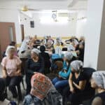 Murshid ladies (who were once unreached), approximately 30, who have already been evangelized sit in a white walled room in charis, some at tables in midst conversation with on another.