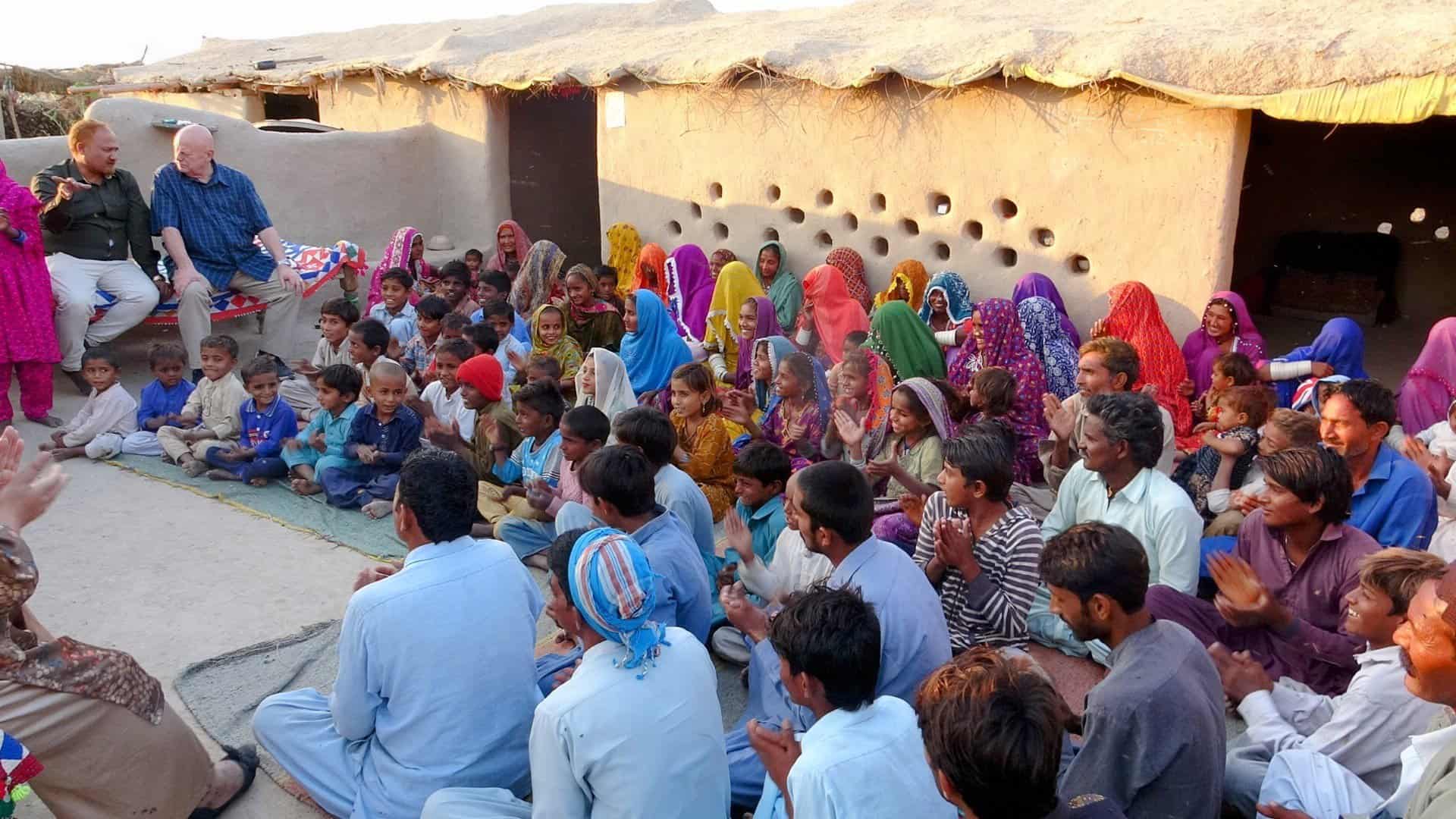 An outdoor gathering of a Pakistani church community in a desert setting, with members of all ages sitting on the ground, joyfully listening to the preached Word of God.