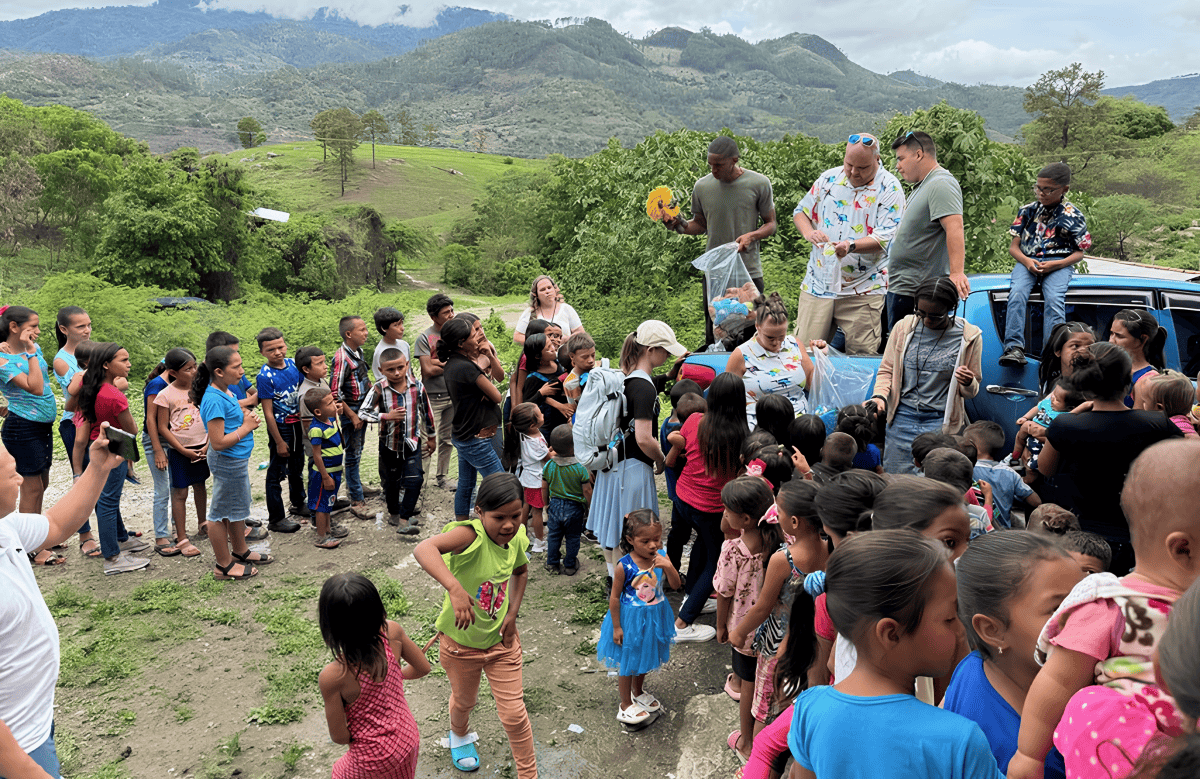 Group of children and adults gathered outdoors in a village in Honduras, with volunteers distributing items from a vehicle.