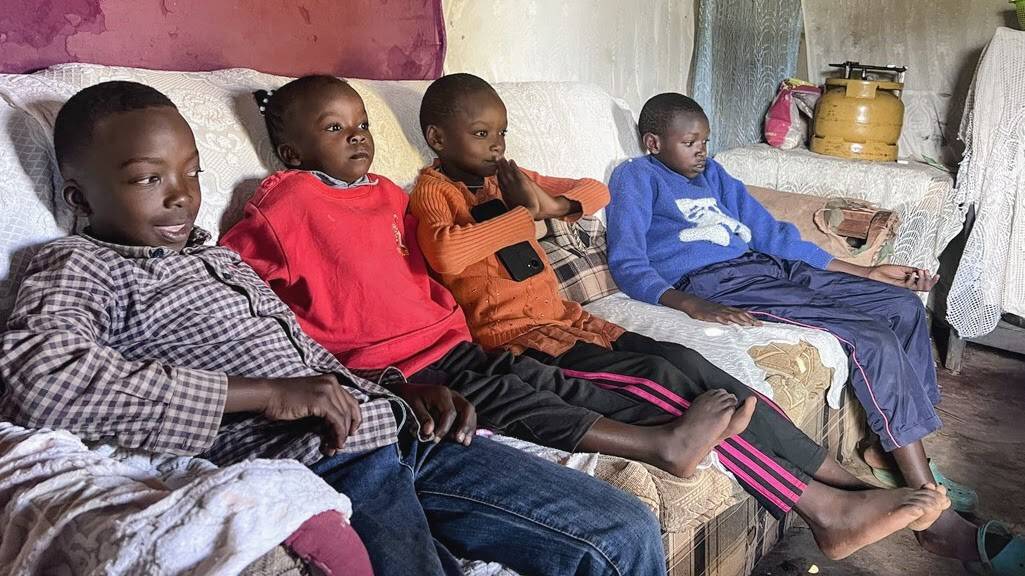 Four children sitting on a couch inside a modest home, watching something attentively.