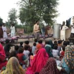 Set in a desert rural village one man is standing preaching infront of a crowd of men, women and children. The crowd are wearing coloring head scarfs, some plain and some with patterns. In the background are several trees as well as 3 foot tall brick walls.