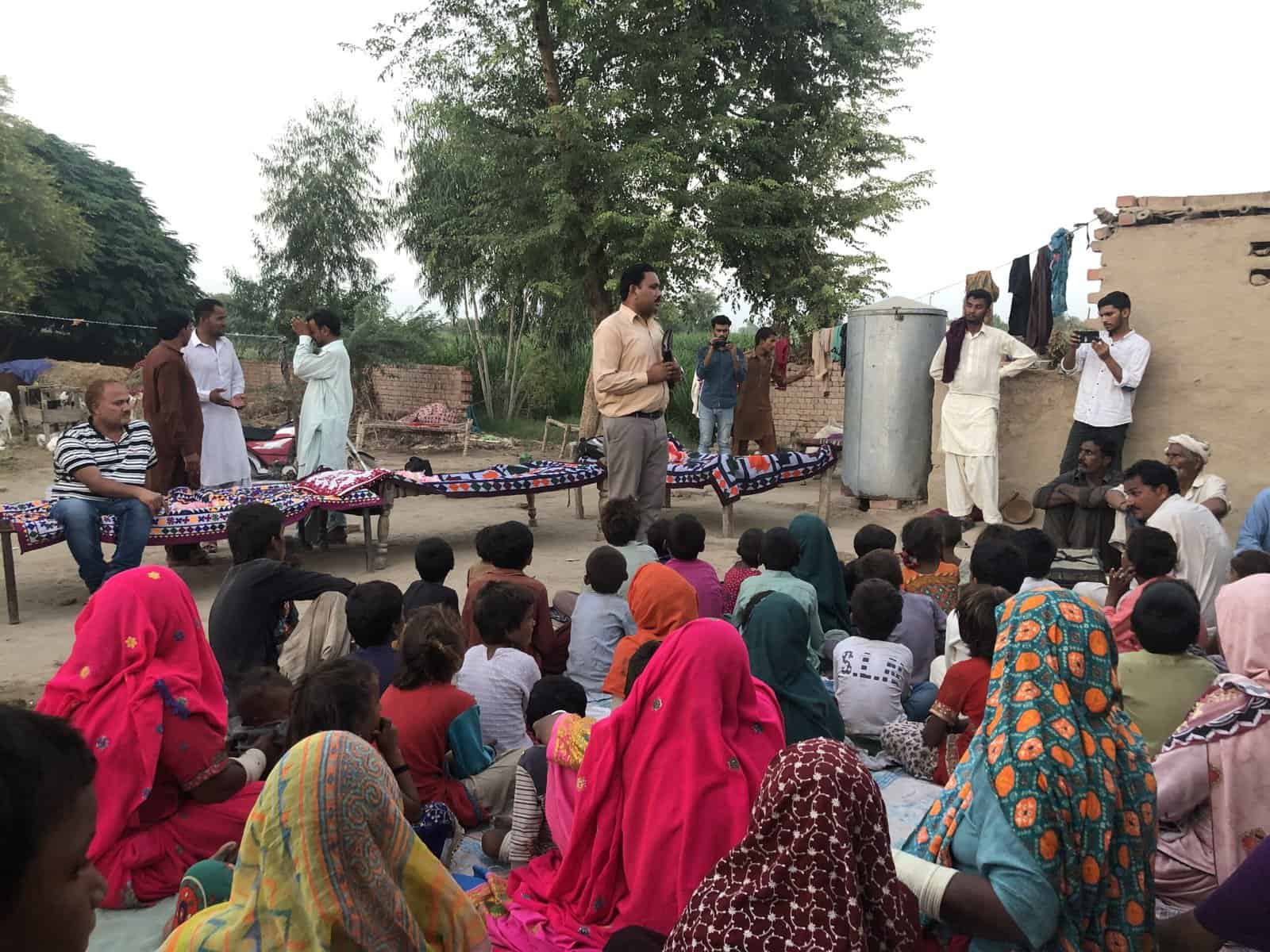 Set in a desert rural village one man is standing preaching infront of a crowd of men, women and children. The crowd are wearing coloring head scarfs, some plain and some with patterns. In the background are several trees as well as 3 foot tall brick walls.