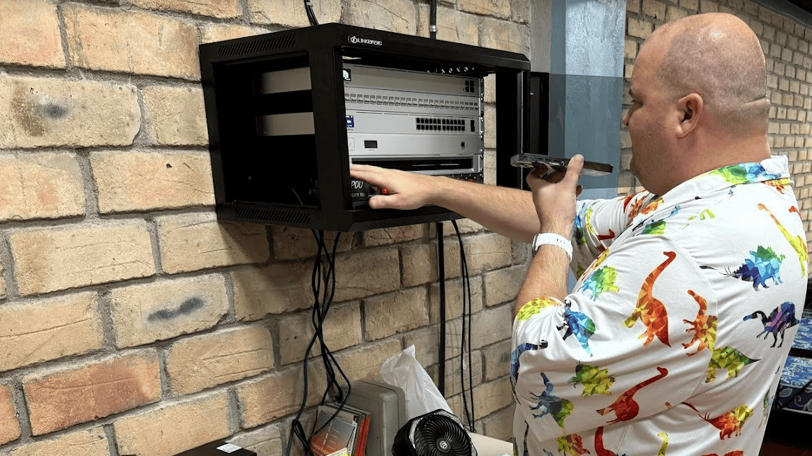 A man wearing a colorful dinosaur-patterned shirt works on installing a Starlink system in a wall-mounted rack at a facility in Honduras.