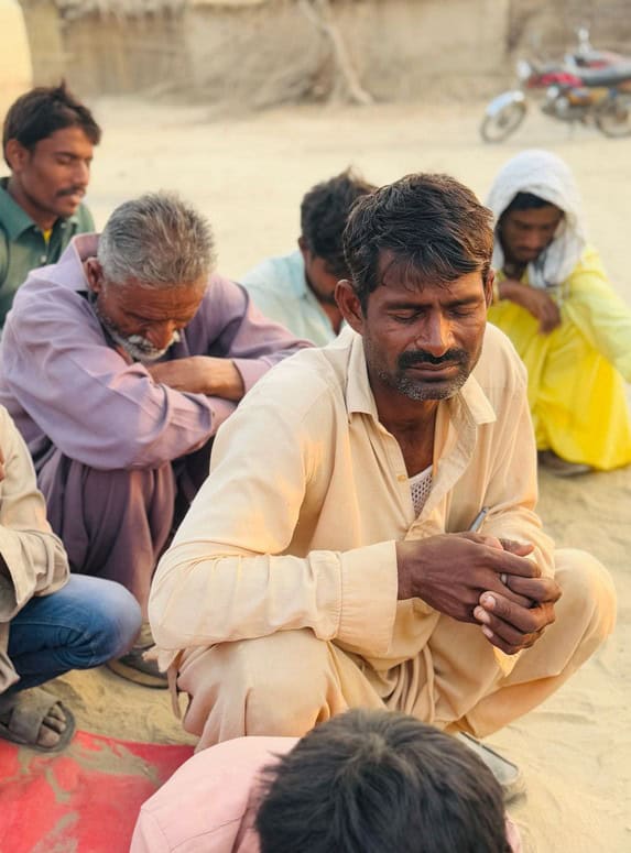 A man has his eyes closed while squating on the sand. Around him are several other people, women and men, who are also sitting with their eyes closed as they pray. In the background you see the side of a sand hut building and a motorcycle.